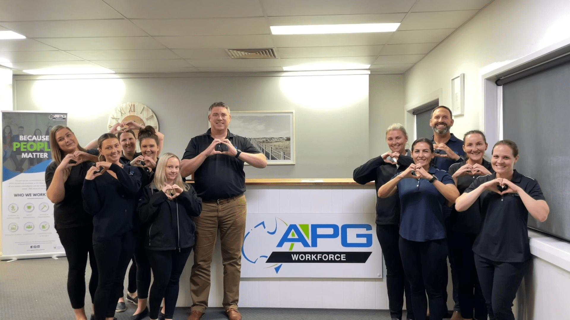 APG Workforce staff in front of APG sign with hands symbolising a heart